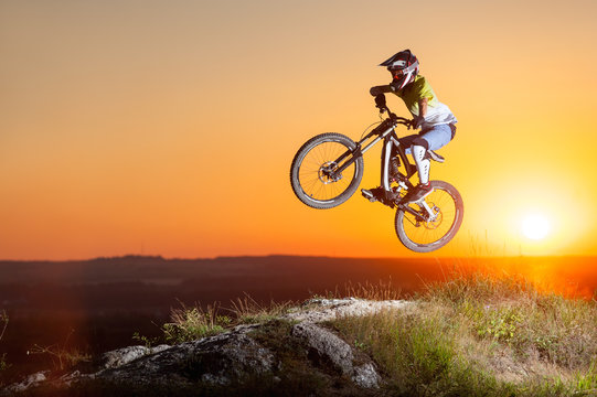Sunset. Biker flying on a mountain bike on the precipice of hill against evening sky with bright sun. Cyclist is wearing sportswear helmet and glasses. Extreme sport