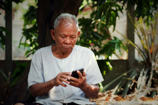 Asian Old Man Using Smartphone Under Big Tree In Old Home.