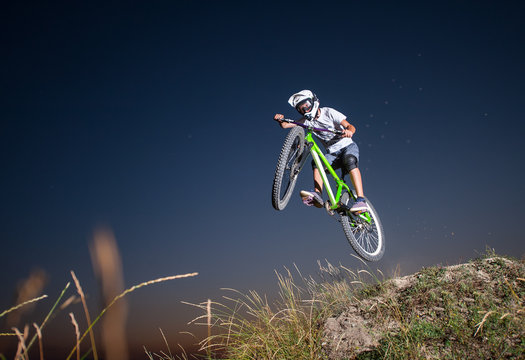 Young guy making high jump on a mountain bike on the hill against evening sky. Cyclist is wearing sportswear helmet and glasses. Bottom view. Extreme downhill.