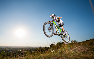 Fototapeta na wymiar Athlete biker jumps on a mountain bike on the hill against blue sky into the distance in the evening. Cyclist is wearing white sportswear helmet and glasses.
