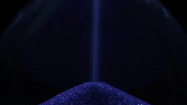 Blue Sand falling in hourglass on a dark background. Extreme close-up.