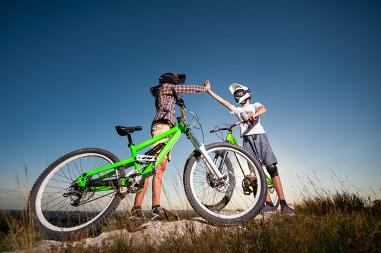 Two male bikers give high five for each other standing with the mountain bicycles on the slope under blue sky. Cyclists are wearing sportswear helmet and glasses. Bottom view