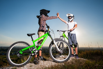 Fototapeta na wymiar Two men give high five for each other standing with the mountain bicycles on the hill under blue sky. Cyclists are wearing sportswear helmet and glasses. Wide angle view