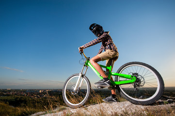 Young cyclist in helmet and glasses on mountain bicycle stands on the precipice of hill under blue sky. Biker is getting ready to ride downhill. Wide angle view