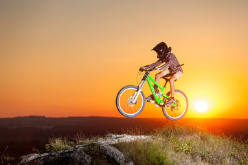 Fototapeta na wymiar Guy high jump on a mountain bike on the top of hill against evening sky with bright sun. Cyclist is wearing sportswear helmet and glasses. Extreme freeride. Sunset