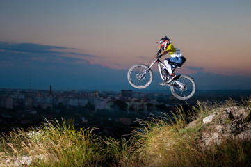 Obraz premium Young guy in helmet and glasses high jumping on a mountain bike on the slope against evening sky and small town into the distance. Downhill cycling.