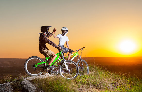 Guys in helmets and glasses stay on the bicycles at the precipice of hill and give high five for each other against evening sky with bright sun at the sunset. Blurred background