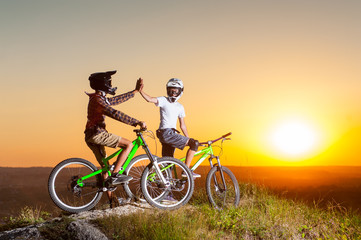 Obraz premium Biker in helmets and glasses stay on the mountain bikes at the precipice of hill and give high five for each other against evening sky with bright sun at the sunset. Blurred background