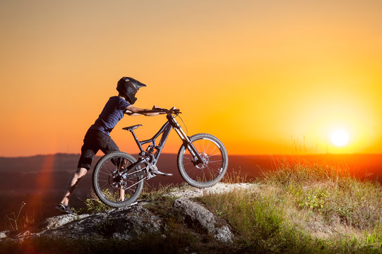 Male cyclist in helmets with mountain bike climbing up on the top of the hill against evening sky with bright sun at the sunset