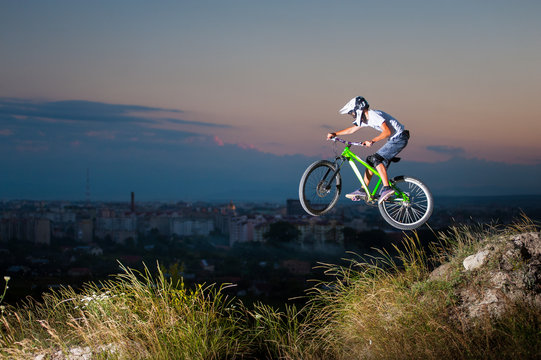 Rider in helmet and glasses flying on a mountain bike on the hill against evening sky and small town into the distance. Extreme downhill cycling.