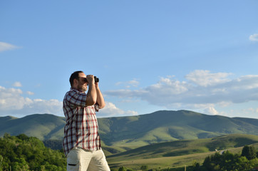 Man is using binoculars to watch divine summer scenery of green forest and grass on surrounding hills and mountain