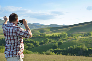 Man is using binoculars to watch wonderful summer scenery of green forest and grass on surrounding hills and mountain