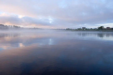 Morning nature scene: fog reflected in the water surface along with reeds, pink and blue sky, trees and a home on the shore.
