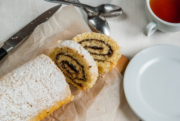Biscuit roll with chocolate cream inside and powdered sugar on top. Roll is sliced, the next tea cup and plate with one slice of dessert. On a white wooden table, top view