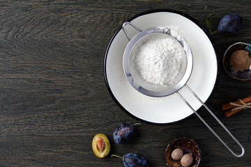 Plum pie cooking process. Powdered sugar, plum fruits and spices on dark wooden background.
