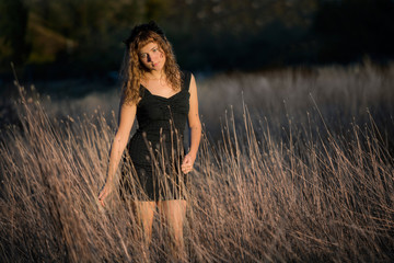 Young female standing in tall dry hay grass in black dress and red hair looking down sad © Andriy Blokhin