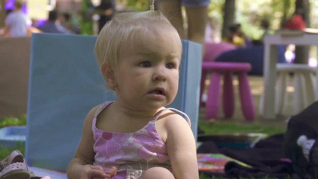 Little girl baby surprise stunned looks into the distance while sitting on the grass in a city park, slow motion
