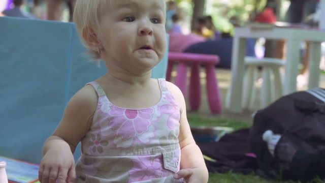 Little girl grimaces and makes faces sitting on the grass in the park, slow motion