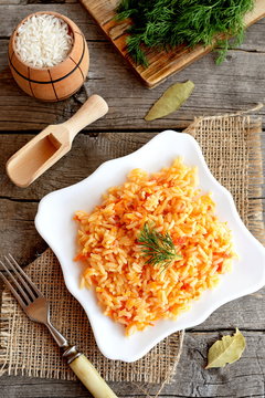 Vegetable risotto on a plate, fork, green dill, cutting board on old wooden background. Rice cooked with tomatoes, carrots, garlic and spices. Easy vegetarian risotto recipe