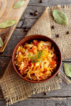 Spicy braised cabbage in a bowl and on old wooden background. Cabbage braised with carrots, tomatoes and garlic and garnished with a sprig of dill. Vegetable recipe