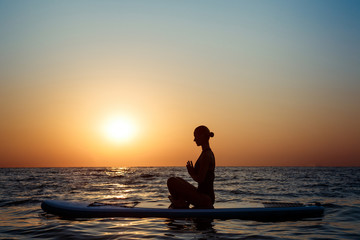 Silhouette of beautiful girl practicing yoga on surfboard at sunrise.
