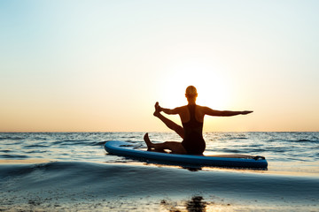 Fototapeta premium Silhouette of beautiful girl practicing yoga on surfboard at sunrise.