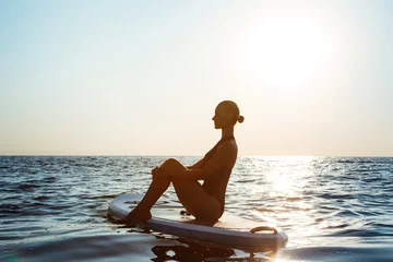 Fotobehang Silhouette of beautiful girl practicing yoga on surfboard at sunrise. © Cookie Studio