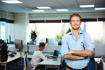 Young successful businessman smiling, posing with crossed arms, over office background.