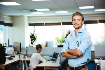 Naklejka na ściany i meble Young successful businessman smiling, posing with crossed arms, over office background.