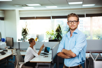 Young successful businessman smiling, posing with crossed arms, over office background.
