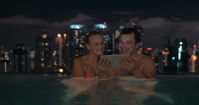 Night view of young beautiful couple using tablet and kissing in swimming pool on the roof of skyscraper on foreground and modern city landscape on the background. Kuala Lumpur, Malaysia