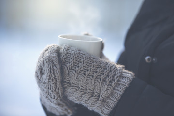 A person is holding a cup of hot drink outdoor. The weather is very cold and the mug is smoking. The person has a jacket and wool mittens in her hands. 