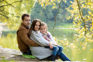 Naklejka na ściany i meble Young lovely family sitting at the lake shore in the park and enjoying warm sunny weather. Looking at the camera with sweet smiles. Happy healthy family lifestyle.