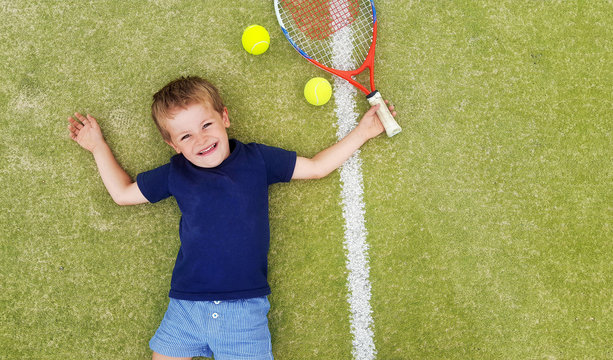 A young blond boy smiling and laying on a tennis court, with racket and balls.