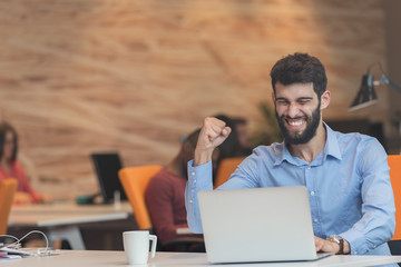 Young bearded caucasian modern business man sitting in a startup office using laptop, looking downward the screen, smiling - business, work, technology concept