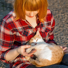 Redhead girl in a plaid shirt holding a cat in her arms, a cat curled up and looking at the camera