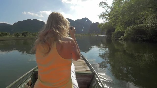 Excursion in Halong bay, boat island tours. View of woman on boat taking pictures by reflex camera on the river, hills and trees on the background. Hanoi, Vietnam