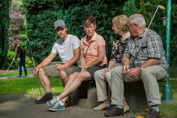 Drei Generationen beim Minigolf. Grosseltern, Tochter und Enkel pausieren auf der Bank.