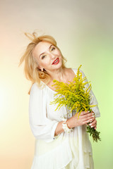 Взрослая женщина в белой блузе мило улыбается и держит желтые цветы.