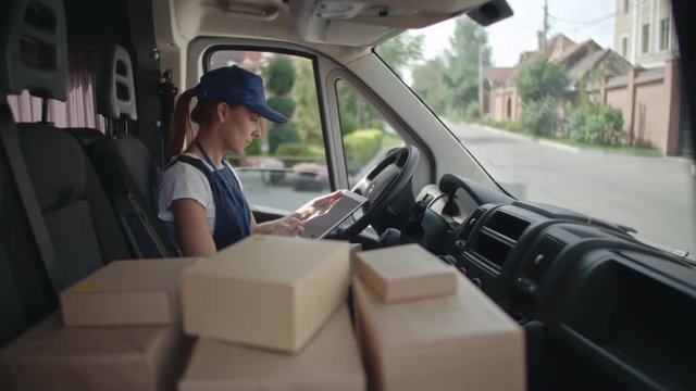 Delivery woman in shipping uniform sitting in van, preparing packages and using her digital tablet to fill in application form