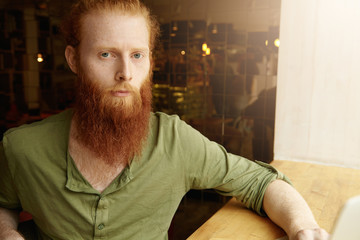 Portrait of young redhead freelancer dressed casually, looking at camera with serious and thoughtful expression, using wireless Internet connection for distant work, sitting in front of his laptop