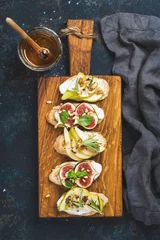 Fototapeten Crostini with pear, ricotta cheese, honey, figs, nuts and herbs. Breakfast toasts or snack sandwiches on rustic wooden board over dark blue grunge plywood background. Top view, vertical composition © sonyakamoz