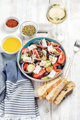 Greek salad with olive oil, bread, oregano, pepper and glass of white wine over old white painted wooden board, top view, selective focus, vertical composition