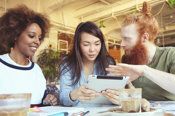Young people working together sitting at coworking space at table with working accessories. Caucasian redhead man with hipster hairstyle presenting brand new vision of business concept using touch pad
