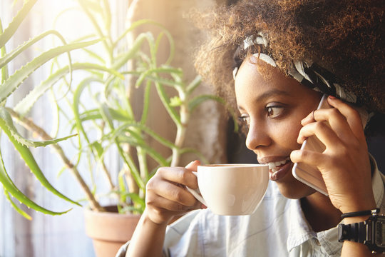 Portrait of thoughtful black woman with Afro haircut, wearing trendy clothes resting at home, looking through window, holding cup of coffee while talking on cell phone, inviting friends to her place