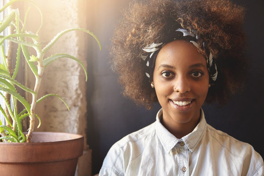 Fashionable dark-skinned hipster girl with Afro haircut wearing denim shirt and do-rag, looking and smiling at camera with happy expression. Cheerful black woman with facial piercing resting at home