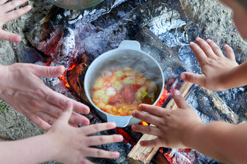 cooking on fire at picnic, hands around food prepared in kettle on wood, potatoes and tomatoes, healthy vegetarian food