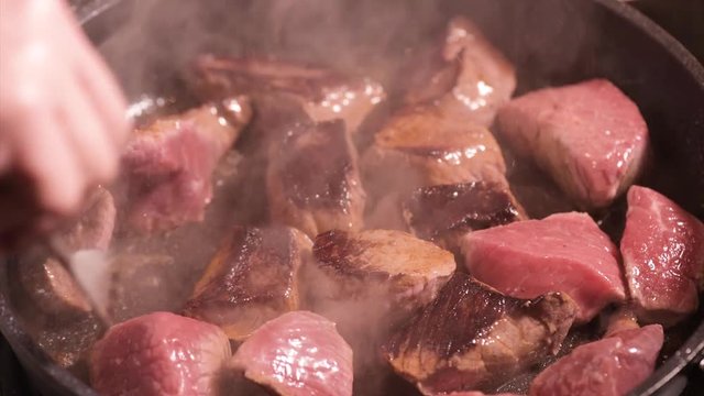 Frying beef sirloin in a hot pan. Turning pieces of  beef meat - close-up on top of a kitchen tile. Cook with fork preparing meat.