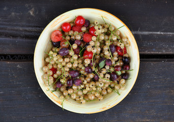 Fresh berries: white currant, raspberry, gooseberry, cherry in a round ceramic plate on dark wooden surface. Top view close up.