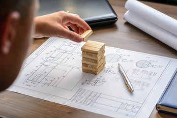 The man (businessman, architect) draws a plan, graph, design on large sheet of paper at office desk and builds model house from wooden blocks 